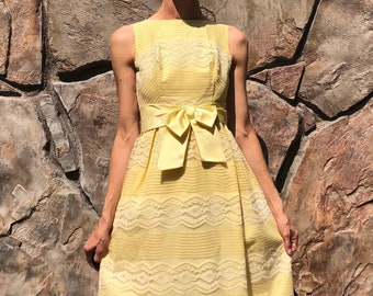 Puede incluir: Un vestido maxi amarillo con una superposición de encaje blanco y una faja amarilla a juego atada en un lazo en la cintura.