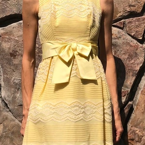 May include: A sleeveless yellow dress with a white lace overlay and a matching yellow satin sash tied in a bow at the waist.