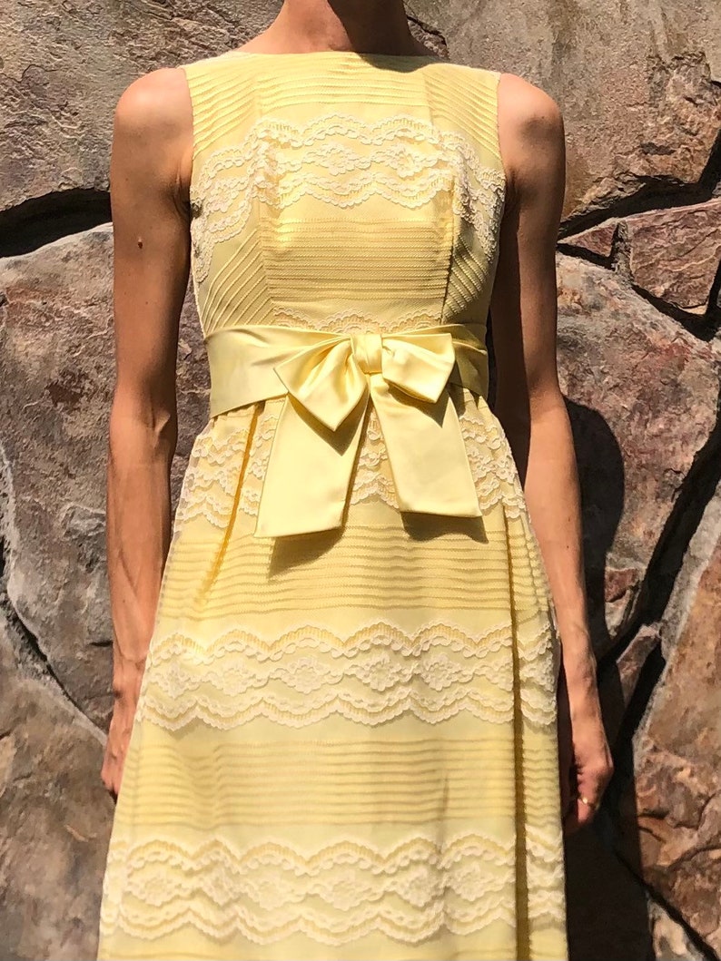 May include: A sleeveless yellow dress with a white lace overlay and a matching yellow satin sash tied in a bow at the waist.