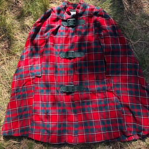 May include: Red and green plaid cape with three black leather buckles. The cape has a high collar and a wide, flowing shape.