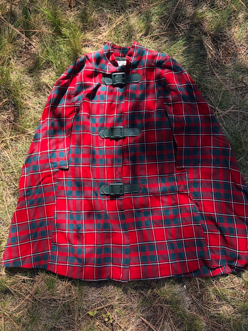 May include: Red and green plaid cape with three black leather buckles. The cape has a high collar and a wide, flowing shape.