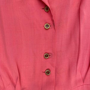 May include: A close-up of a pink button-down shirt with gold buttons. The shirt is made of a smooth, textured fabric.