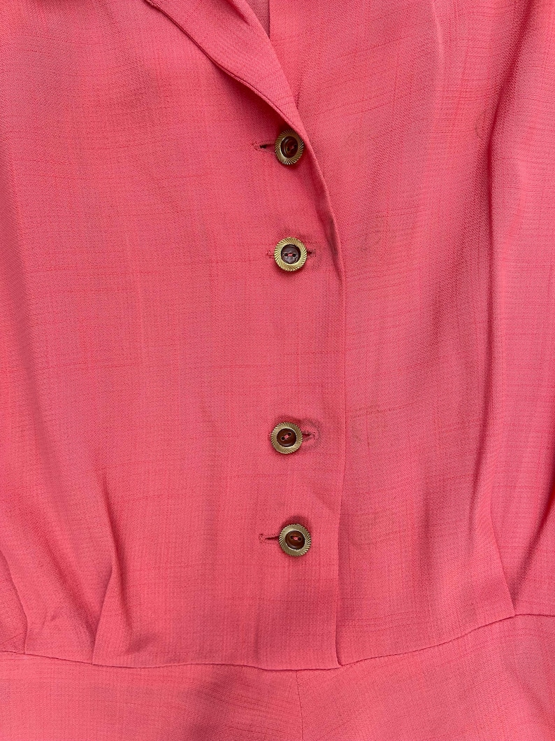 May include: A close-up of a pink button-down shirt with gold buttons. The shirt is made of a smooth, textured fabric.