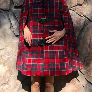May include: A red and black plaid cape with three black leather buckles. The cape has a high collar and is made of a soft, warm fabric.