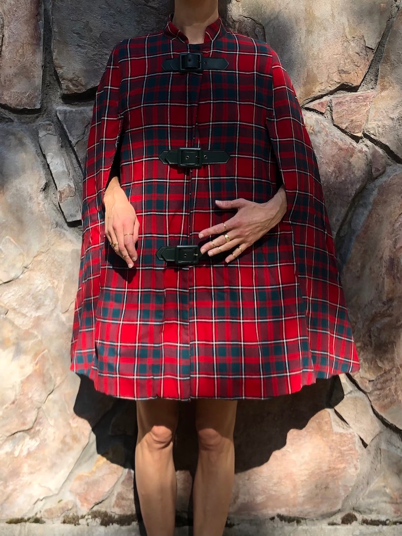 May include: A red and black plaid cape with three black leather buckles. The cape has a high collar and is made of a soft, warm fabric.