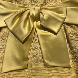 May include: A yellow satin bow tied on a yellow lace dress. The bow is large and the dress has a delicate lace pattern.