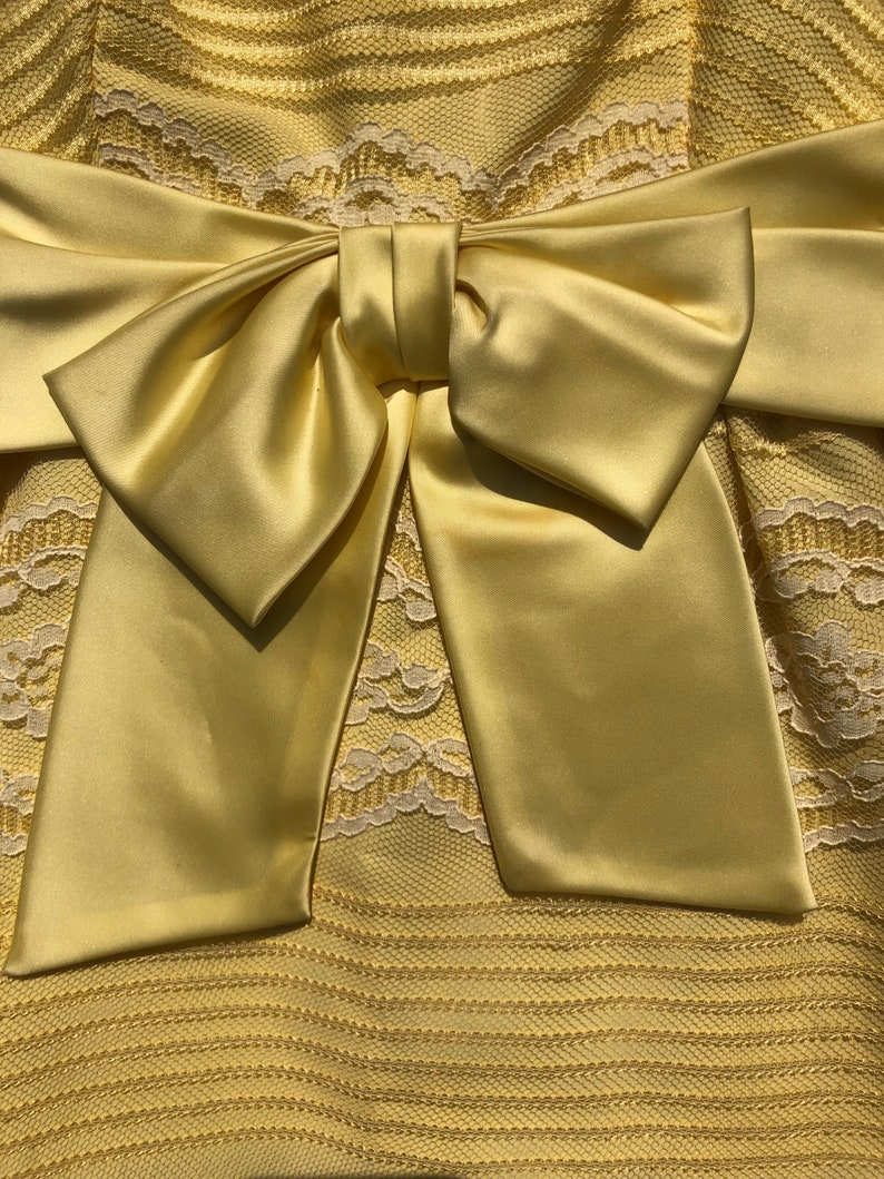 May include: A yellow satin bow tied on a yellow lace dress. The bow is large and the dress has a delicate lace pattern.