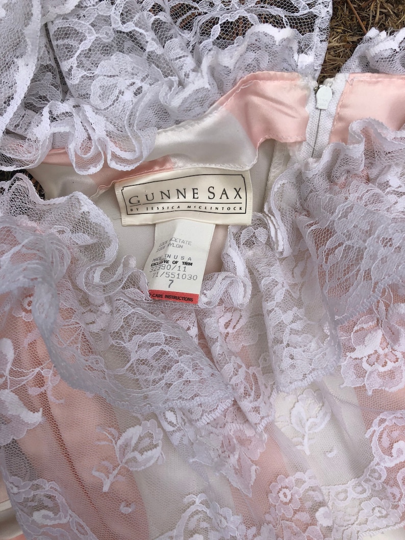 May include: A white lace dress with a pink satin lining. The dress has a label that reads "Gunne Sax by Jessica McClintock" and a size tag that says "Size 7".