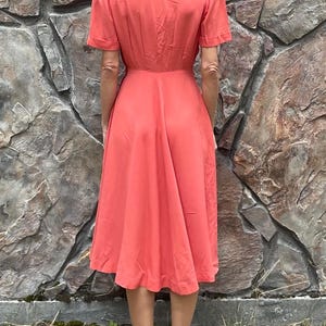 May include: A coral-colored midi dress with a V-neckline and short sleeves. The dress has a fitted bodice and a flowing skirt.