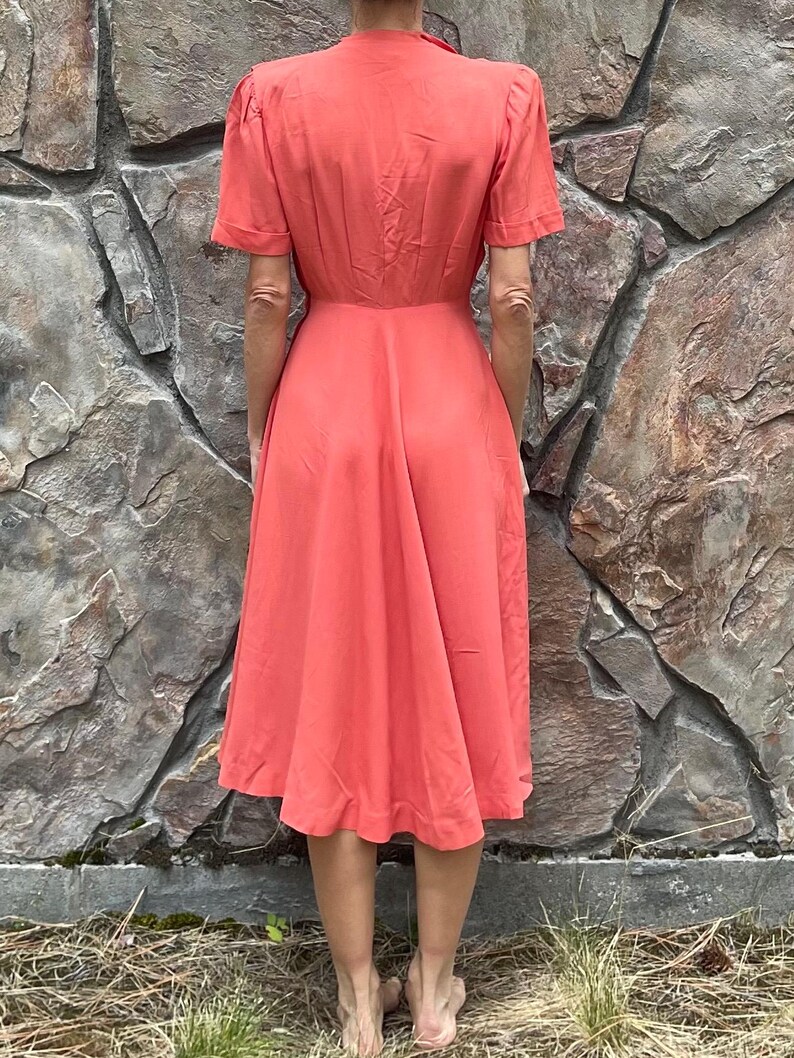May include: A coral-colored midi dress with a V-neckline and short sleeves. The dress has a fitted bodice and a flowing skirt.