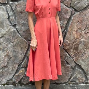 May include: A coral colored midi dress with a collared neckline and short sleeves. The dress has a button-up front and a fitted bodice with a flared skirt.