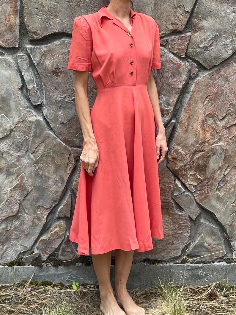 May include: A coral colored midi dress with a collared neckline and short sleeves. The dress has a button-up front and a fitted bodice with a flared skirt.