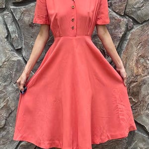 May include: A coral colored vintage dress with a collared neckline and short sleeves. The dress has a fitted bodice and a full skirt.
