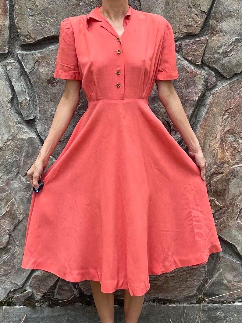 May include: A coral colored vintage dress with a collared neckline and short sleeves. The dress has a fitted bodice and a full skirt.