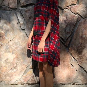 May include: A red and black plaid cape with a high collar and black buckle closures. The cape is made of a soft, lightweight fabric and has a flowing silhouette.