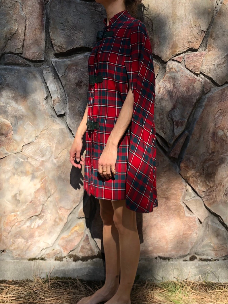 May include: A red and black plaid cape with a high collar and black buckle closures. The cape is made of a soft, lightweight fabric and has a flowing silhouette.