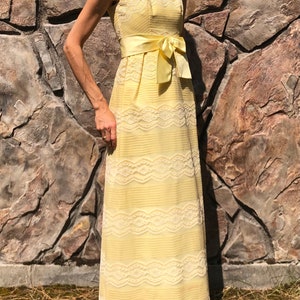 May include: A pale yellow maxi dress with a white lace overlay and a matching satin sash tied in a bow at the waist.