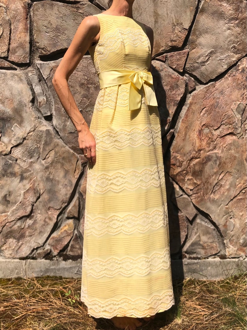 May include: A pale yellow maxi dress with a white lace overlay and a matching satin sash tied in a bow at the waist.