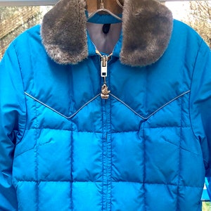 May include: A blue puffer jacket with a faux fur collar. The jacket has a zipper closure and a silver zipper pull with a bird design.