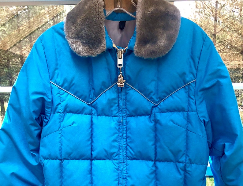 May include: A blue puffer jacket with a faux fur collar. The jacket has a zipper closure and a silver zipper pull with a bird design.