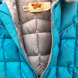 May include: A teal blue quilted jacket with a zipper closure and a fur-lined hood. The jacket has a label that reads "Downright Comfortable Since 1972" and "A Davin Operation, Sioux Falls, South Dakota".