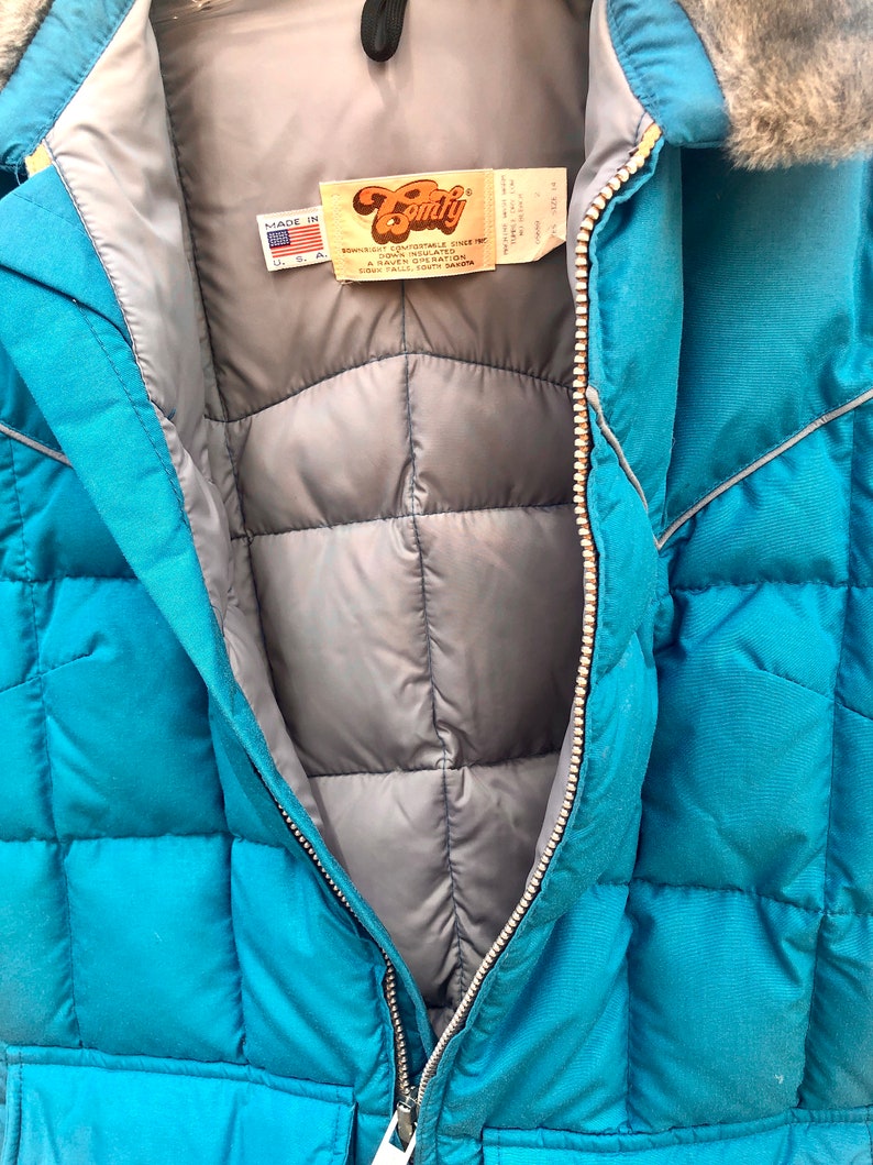 May include: A teal blue quilted jacket with a zipper closure and a fur-lined hood. The jacket has a label that reads "Downright Comfortable Since 1972" and "A Davin Operation, Sioux Falls, South Dakota".