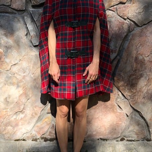 May include: A red and black plaid cape with black leather straps and buckles. The cape is knee-length and has a collar.