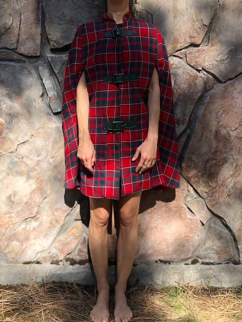 May include: A red and black plaid cape with black leather straps and buckles. The cape is knee-length and has a collar.