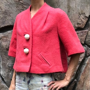 May include: A cropped, pink, fuzzy jacket with three white, round buttons. The jacket has a short sleeve and a single pocket on the left side.
