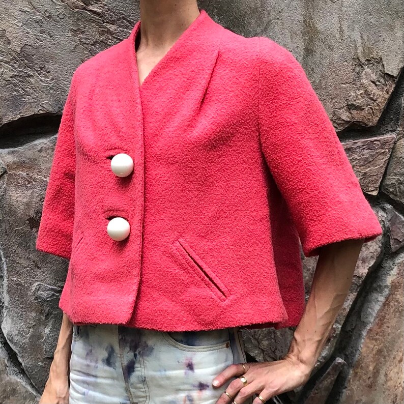 May include: A cropped, pink, fuzzy jacket with three white, round buttons. The jacket has a short sleeve and a single pocket on the left side.