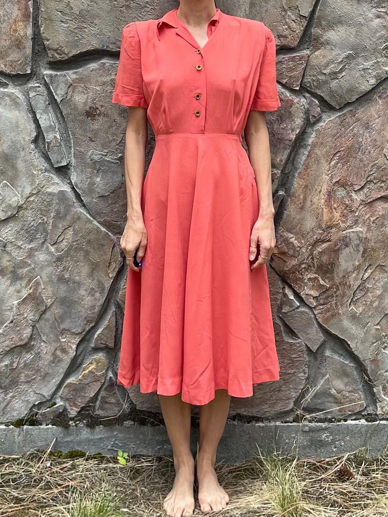 May include: A coral pink vintage dress with a collared neckline, short sleeves, and a button-up front. The dress has a fitted bodice and a full skirt.