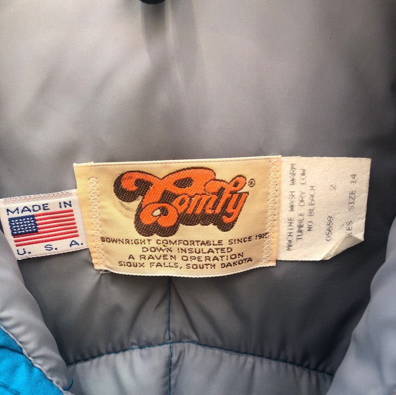 May include: A close-up of a Comfy brand down insulated jacket label. The label is orange and brown with the text "Downright Comfortable Since 1915" and "A Raven Operation, Sioux Falls, South Dakota". The label is attached to a gray jacket with a blue lining.