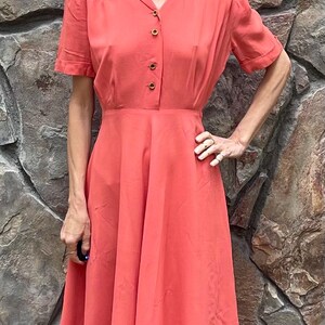 May include: A coral pink midi dress with a collared neckline, short sleeves, and a button-up front. The dress has a fitted bodice and a full skirt.