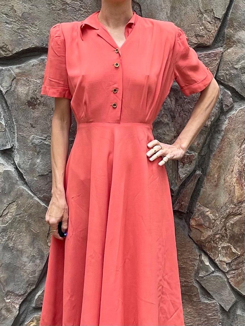 May include: A coral pink midi dress with a collared neckline, short sleeves, and a button-up front. The dress has a fitted bodice and a full skirt.
