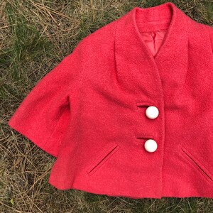 May include: A coral pink short-sleeved jacket with a rounded collar and three white buttons. The jacket has two pockets on the front.