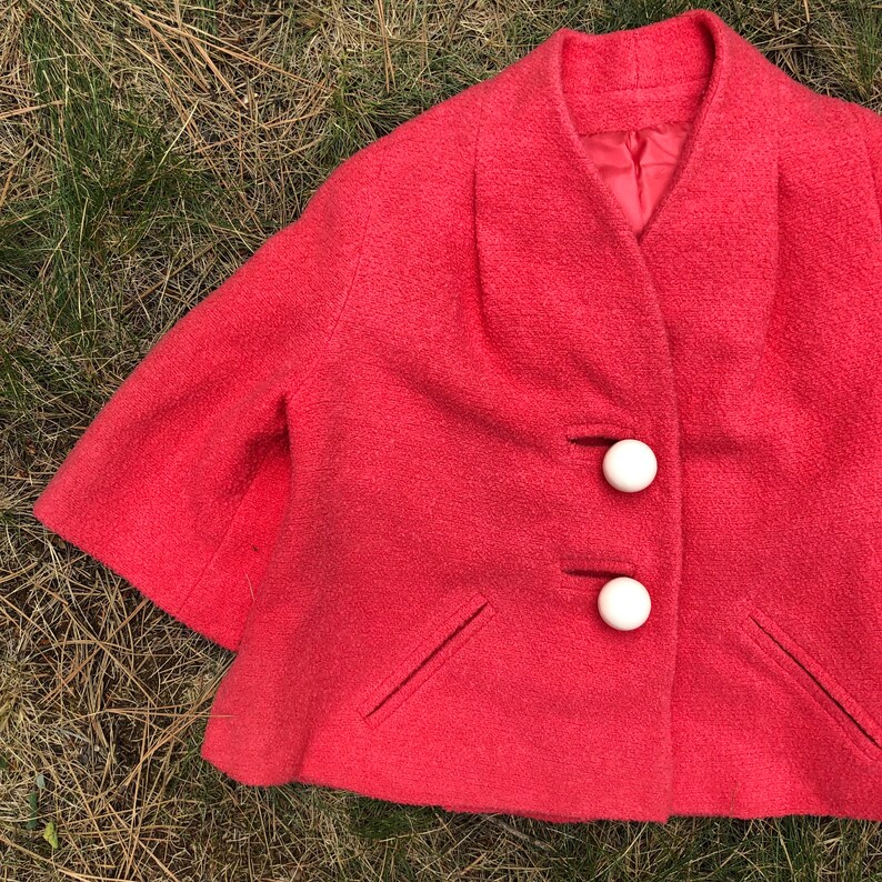 May include: A coral pink short-sleeved jacket with a rounded collar and three white buttons. The jacket has two pockets on the front.