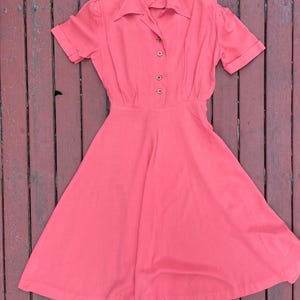 May include: A coral pink vintage dress with a collared neckline and short sleeves. The dress has a button-up front and a full skirt.