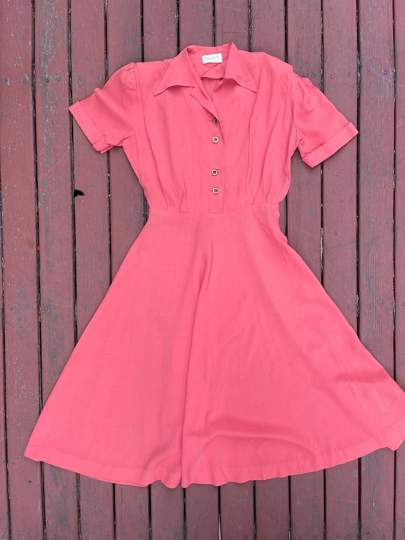 May include: A coral pink vintage dress with a collared neckline and short sleeves. The dress has a button-up front and a full skirt.