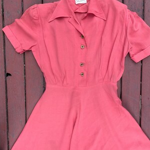 May include: A coral pink vintage dress with a collared neckline and short sleeves. The dress has a button-up front and a full skirt. The label reads "Madeline" in black text.