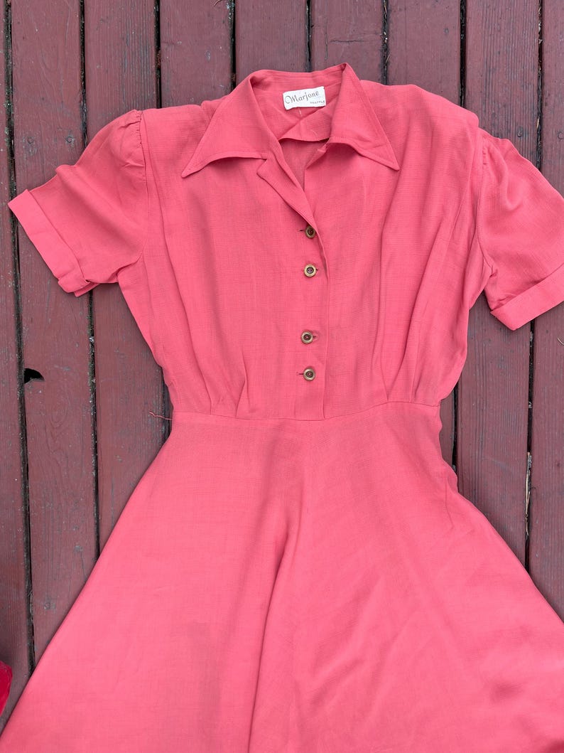 May include: A coral pink vintage dress with a collared neckline and short sleeves. The dress has a button-up front and a full skirt. The label reads "Madeline" in black text.