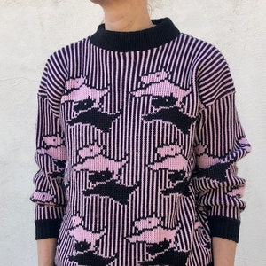 Peut inclure: Un pull rayé noir et rose avec un motif répété d'yeux roses. Le pull a un col rond et des manches longues.