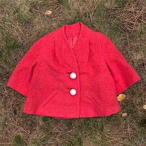 May include: A short, bright pink, vintage jacket with a V-neck and three white buttons. The jacket has a slightly flared shape and two pockets.