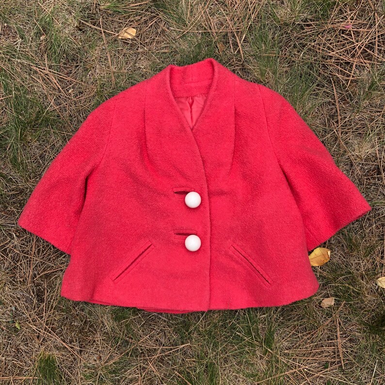 May include: A short, bright pink, vintage jacket with a V-neck and three white buttons. The jacket has a slightly flared shape and two pockets.