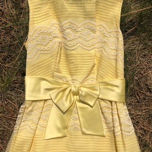 May include: A yellow sleeveless dress with white lace detailing and a matching yellow satin bow at the waist.
