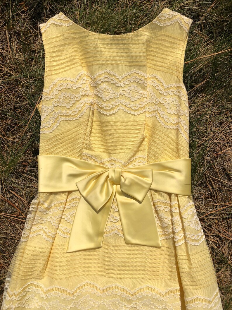 May include: A yellow sleeveless dress with white lace detailing and a matching yellow satin bow at the waist.