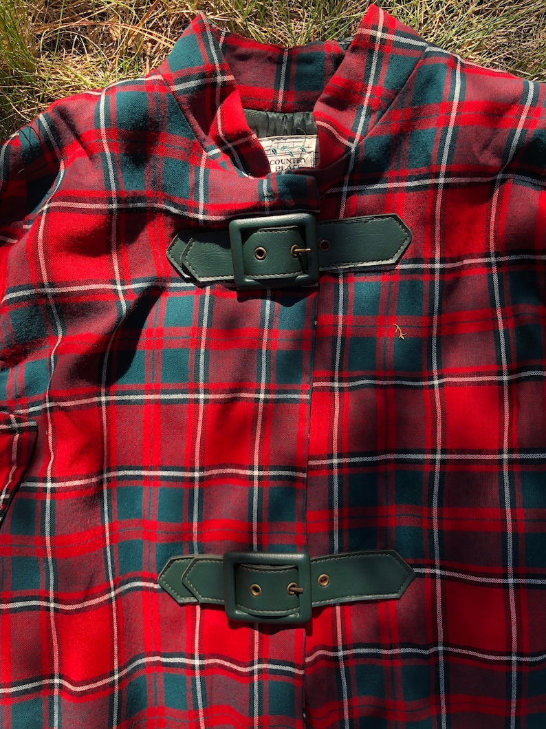 May include: Red and green plaid cape with two leather belts with buckles. The label reads "Country Place".