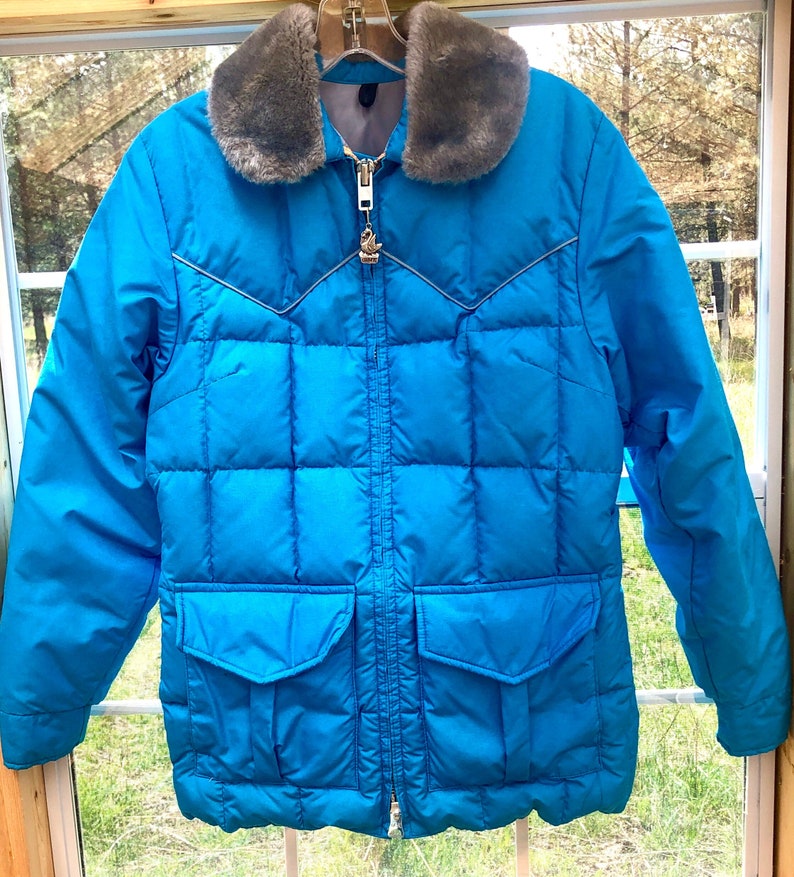 May include: A bright blue puffer jacket with a faux fur collar. The jacket has a zipper closure and a large pocket on the front.