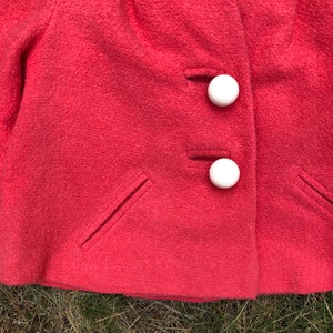May include: A close-up of a pink wool jacket with two white buttons. The jacket has two pockets on the front.