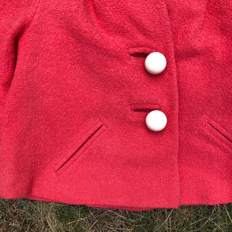May include: A close-up of a pink wool jacket with two white buttons. The jacket has two pockets on the front.
