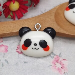 May include: Two white panda bear charms with black eyes and ears, red cheeks, and a silver loop for hanging. The charms are on a wooden spoon and a floral background.
